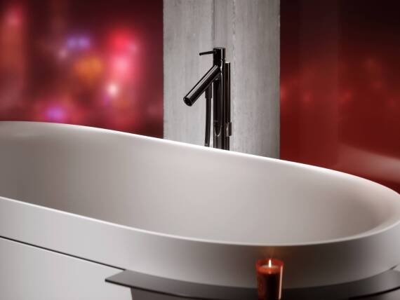 Close-up of an AXOR Starck floor-standing single lever bath faucet in Polished Black Chrome, next to an AXOR Suite Bathtub, against a red-lit background.
