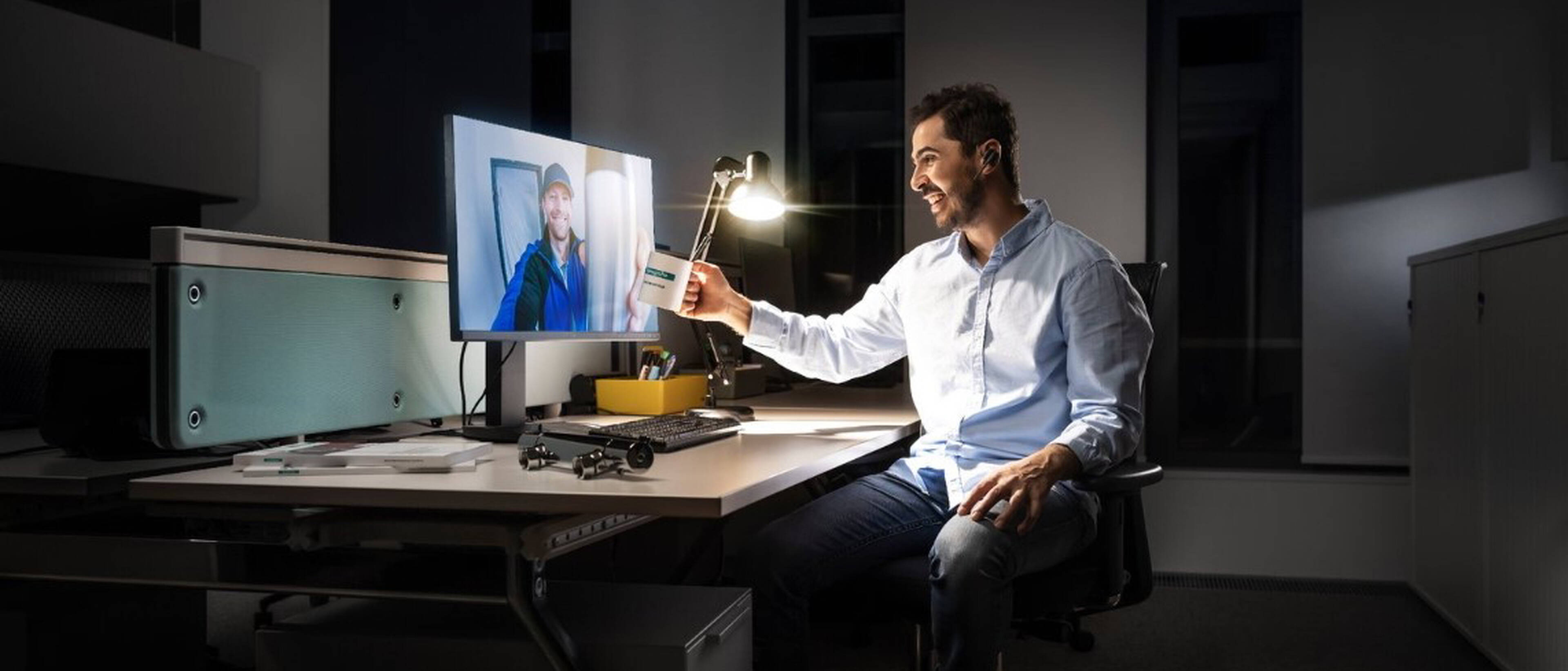 Mann sitzt am Arbeitsplatz und prostet mit seiner Kaffeetasse in Richtung des Bildschirms.