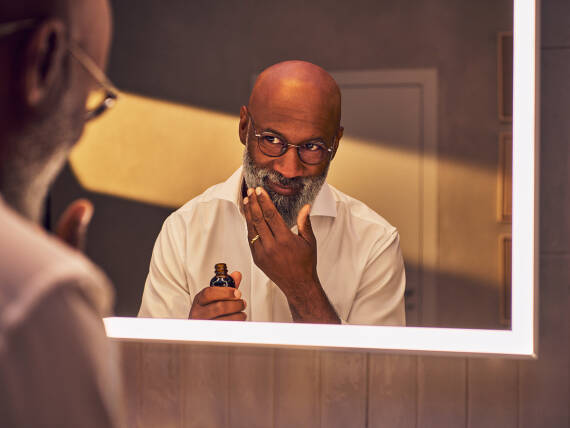Un homme applique de l’huile à barbe devant un miroir éclairé.