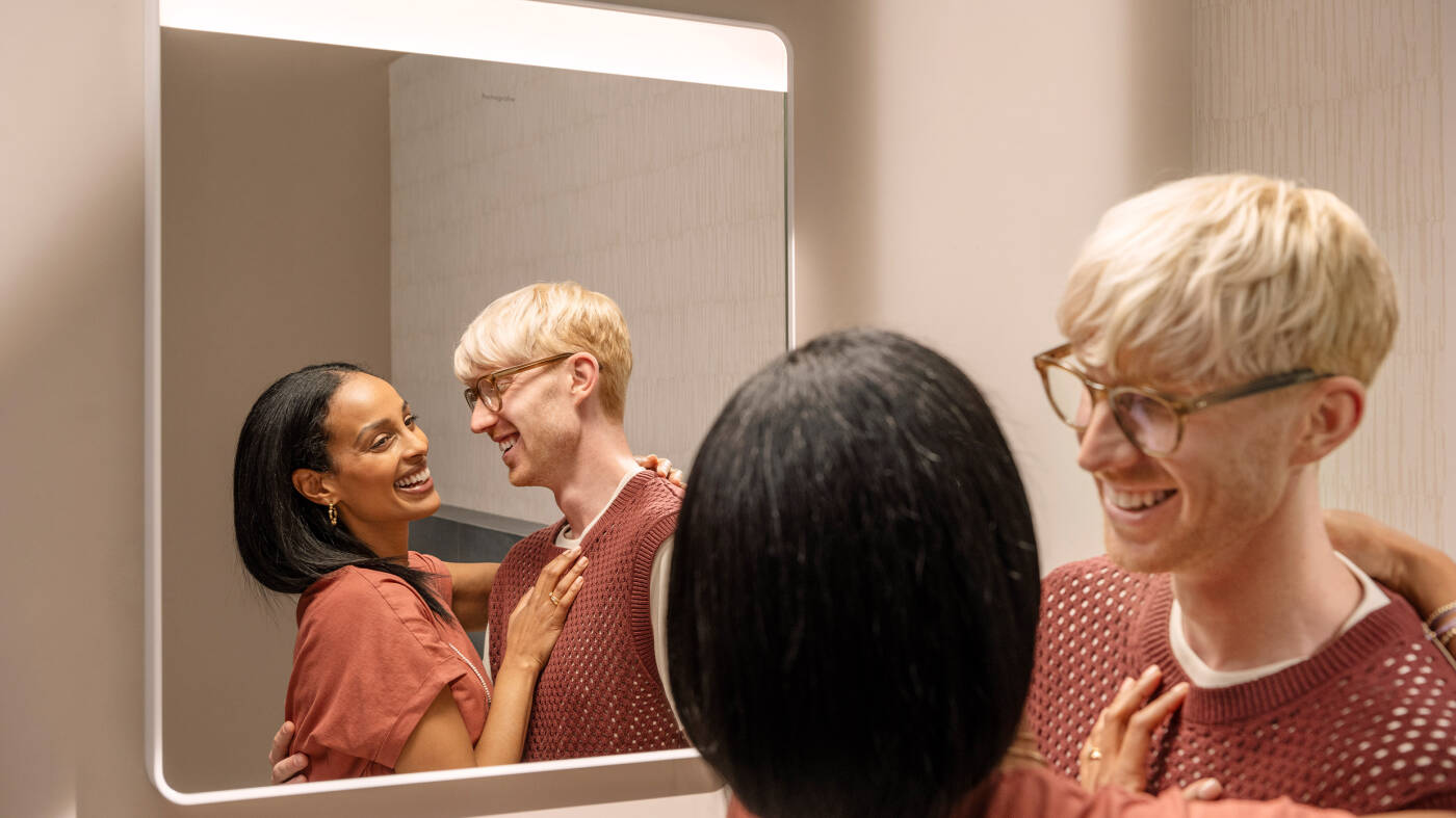 Couple jokes around in front of the lit hansgrohe mirror. 