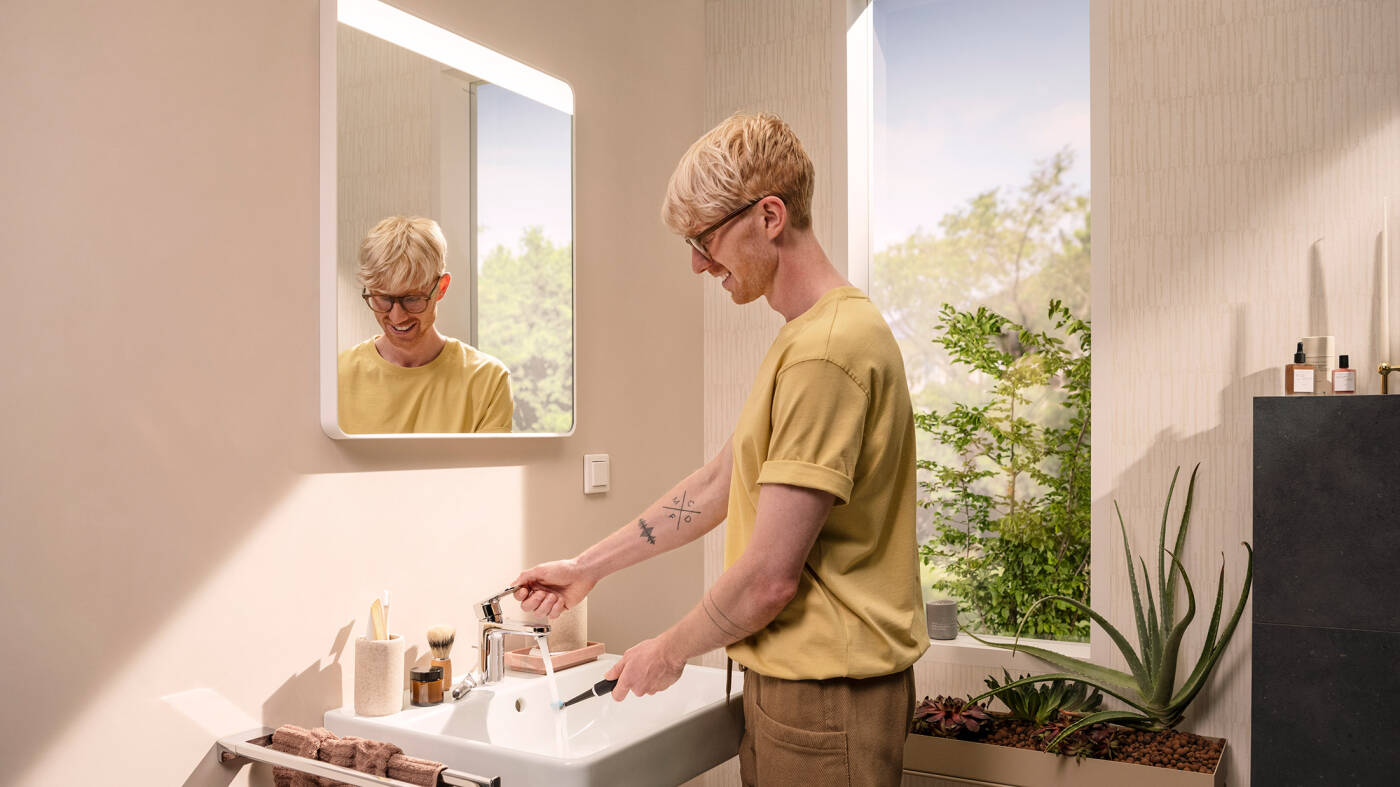 Man operates faucet in front of hansgrohe bathroom mirror with light.