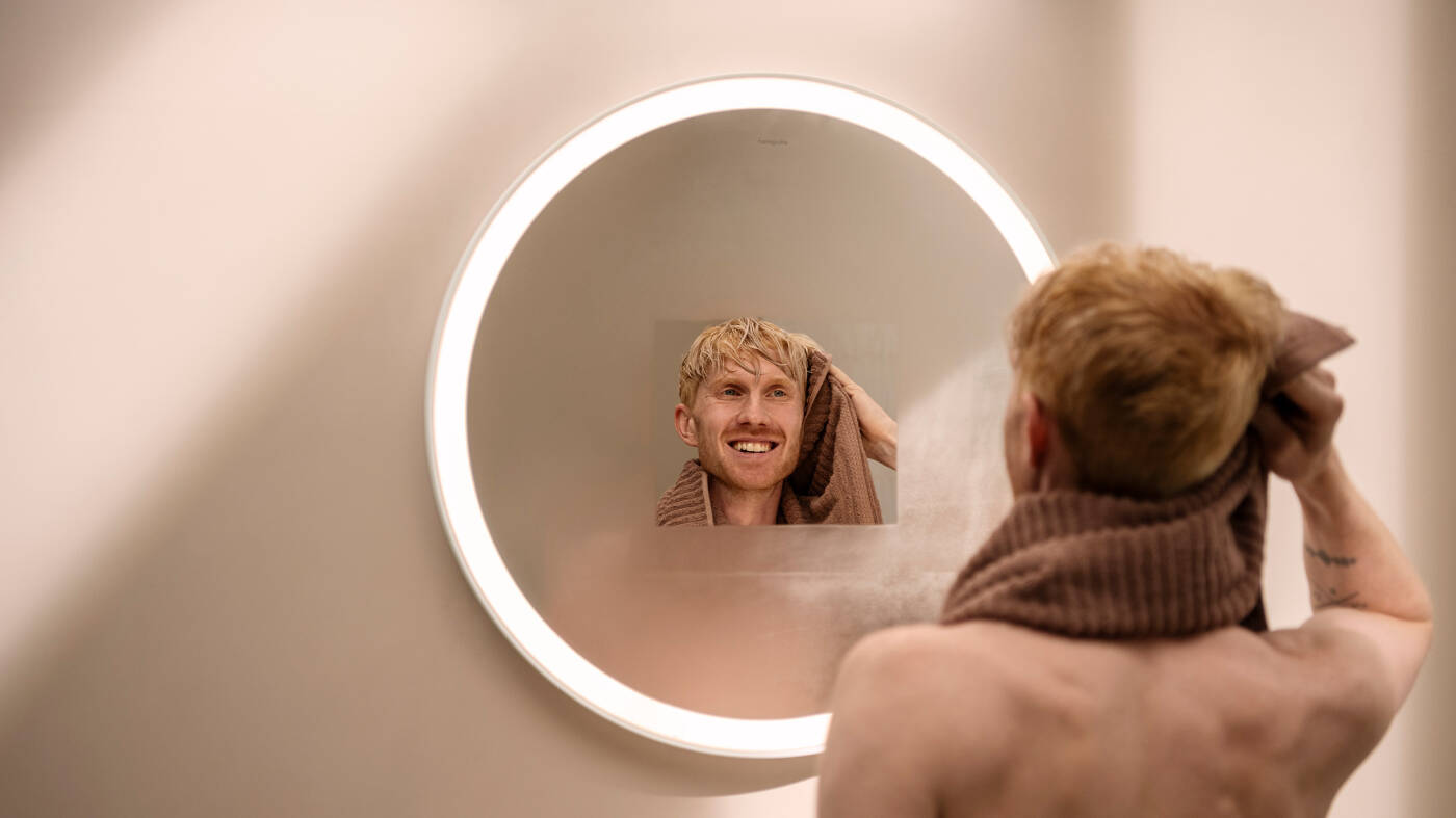 Man stands in front of a round hansgrohe mirror with lighting. 